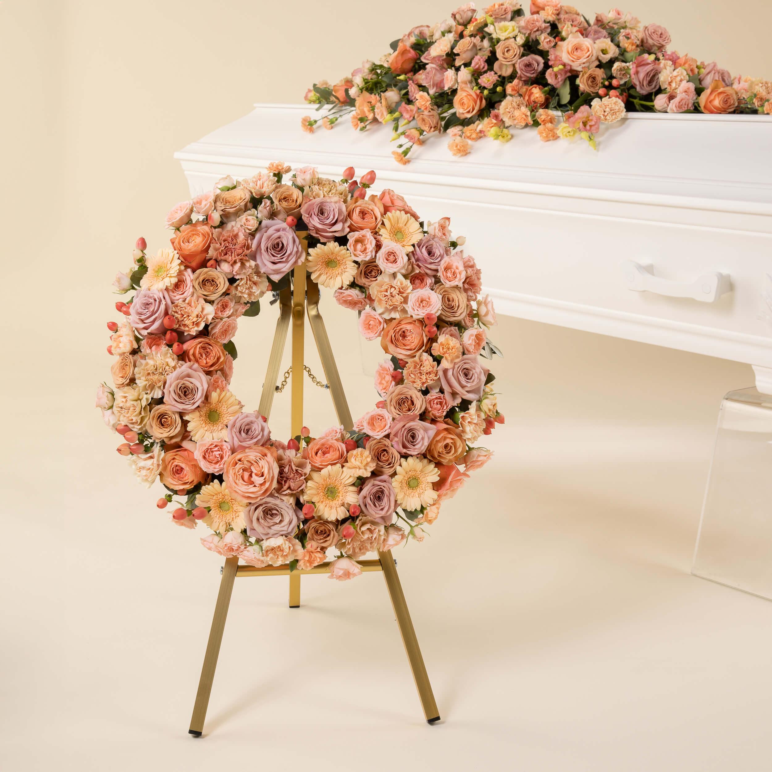Krans til begravelse i duse rosa og ferskenfargede blomster foran hvit kiste med matchende kistedekorasjon.