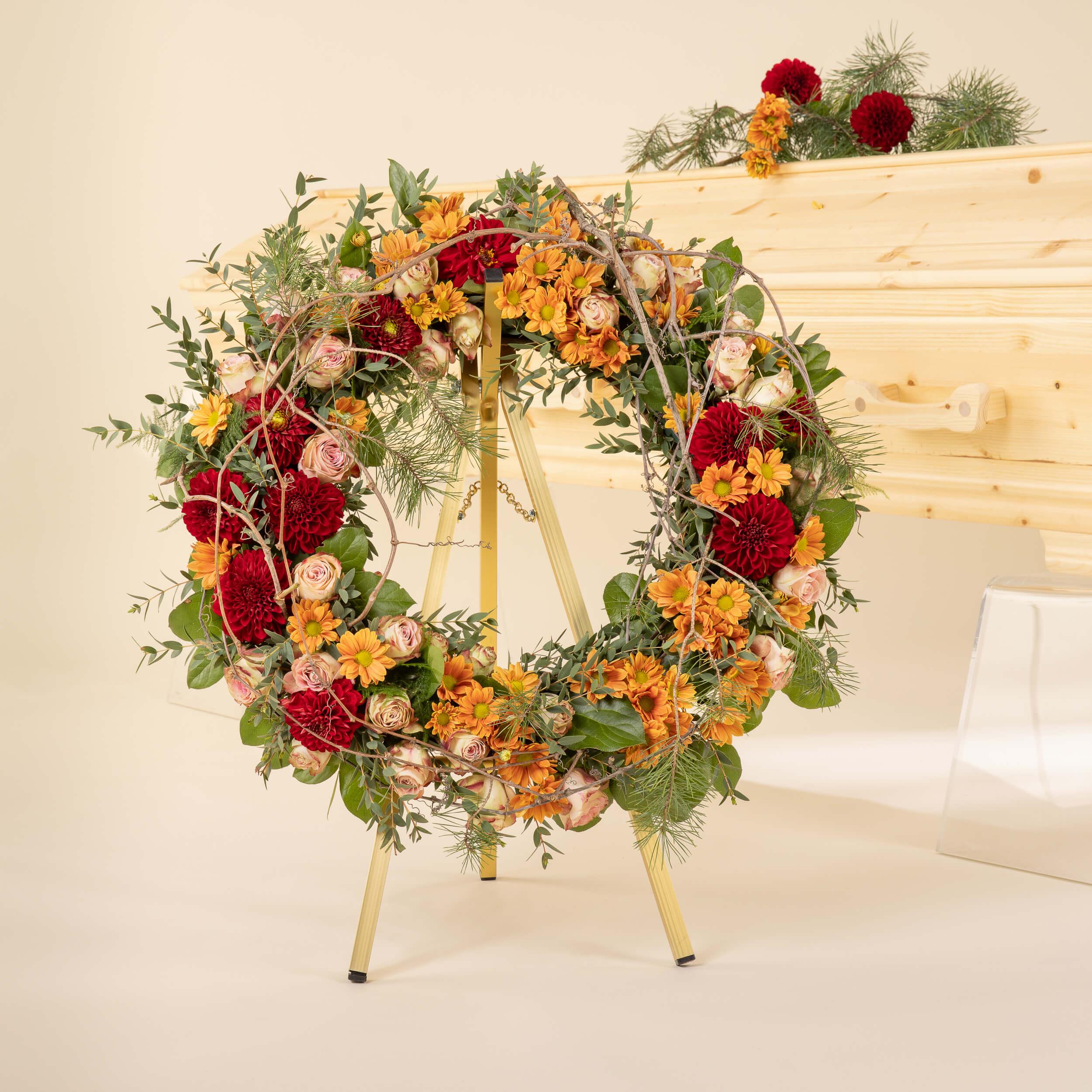 Krans til begravelse med røde og oransje blomster foran kiste i treverk – personlig begravelseskrans i varme og naturlige farger.