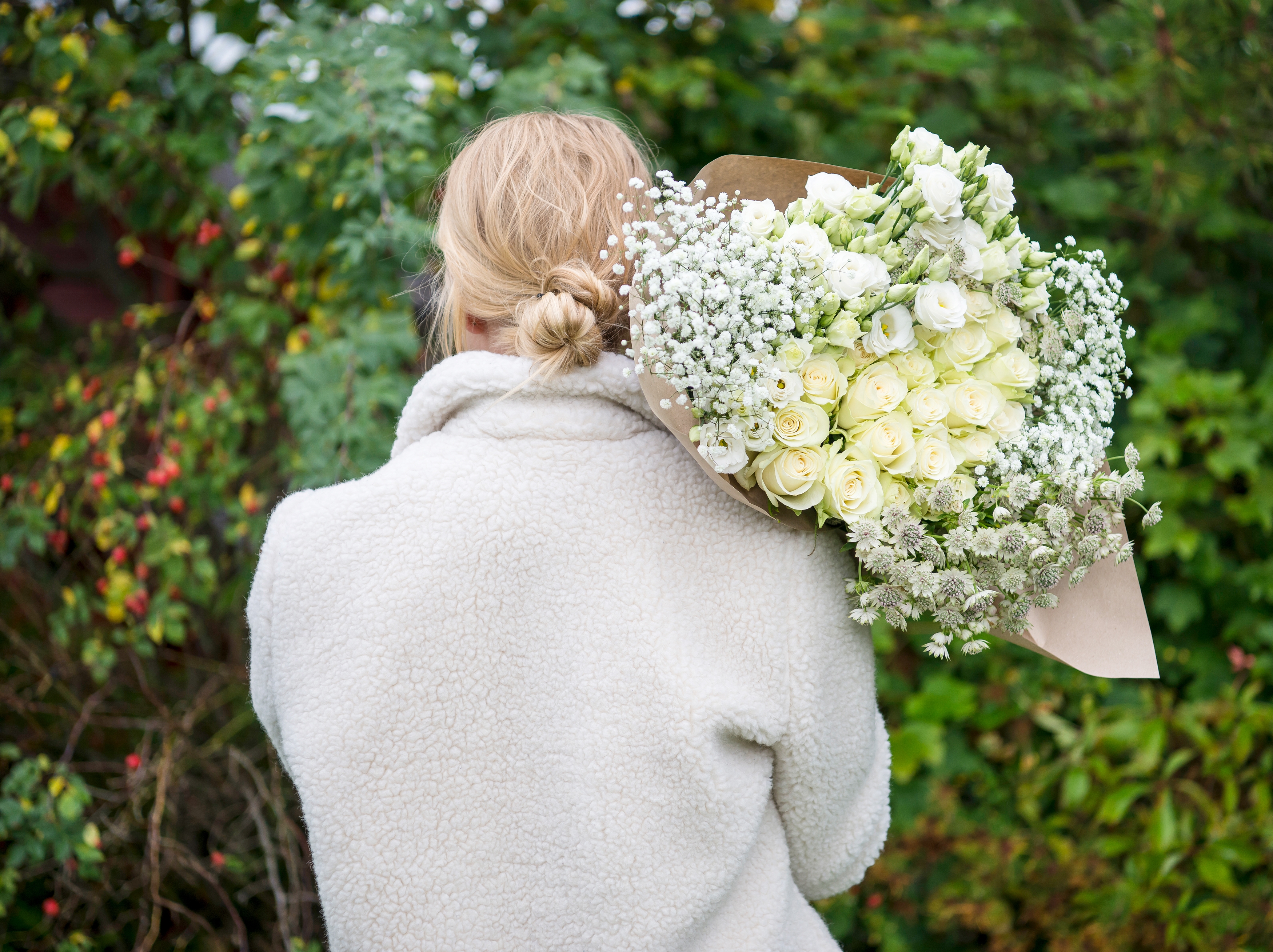 Bukett med hvite roser, hvit lisianthus, hvit brudeslør og hvit astrantia, innpakket med kraftpapir som holdes over skulderen.